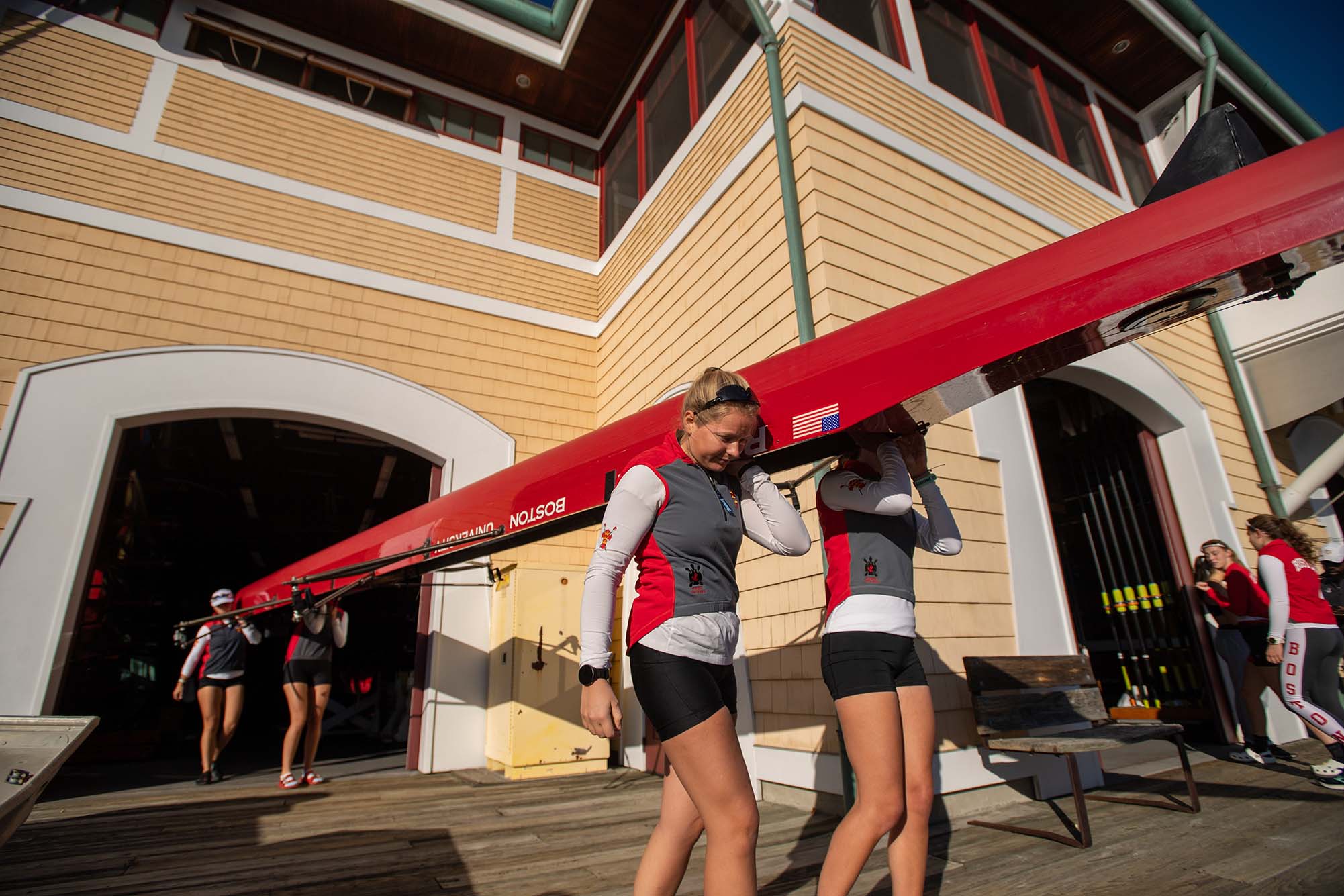 Photo: Athletes from the women’s lightweight crew team head out to practice on the Charles River form the DeWolfe Boathouse Oct 18. The young women wearing BU wet suits hold up a long, red lightweight boat as they head out of a wooden boathouse.