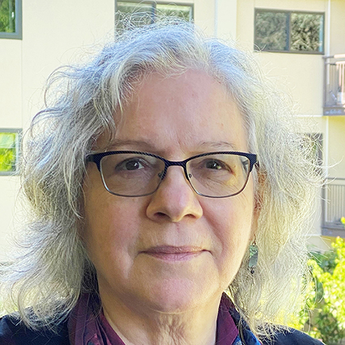 Photo: Headshot of Nancy Harrowitz. An older white woman with shoulder length white hair poses on a bright sunny day in front of a tan building.