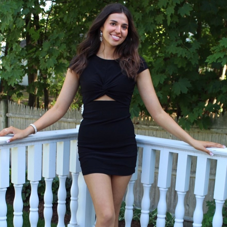 Photo of BU student Sophia Caffrey (COM’25) posing outside. A young white woman wearing a black dress smiles and poses against a white railing behind her.