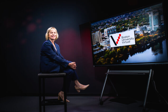 Photo: Wendy Colby, BU's first vice president and associate provost for BU Virtual, poses for a photo in a Questrom online studio. A white woman with shoulder length blonde hair and wearing a navy suit ensemble sits in a chair with hands folded over crossed legs. She sits in a dark room lit with purple backlights as a large monitor to her right displays the Boston University Virtual logo over a stock photo of BU's campus.
