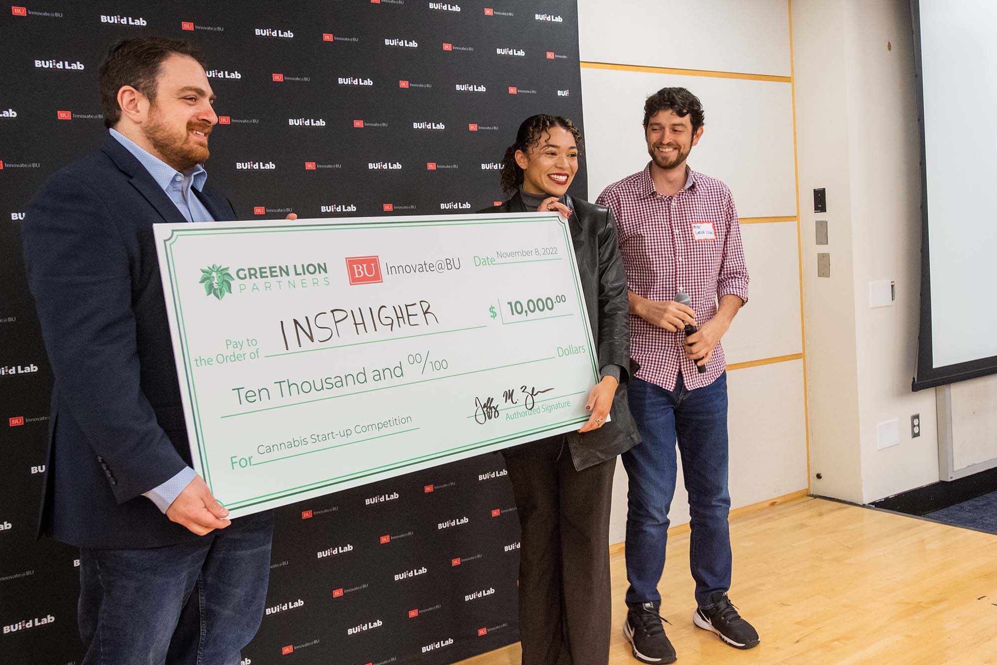 Photo: Green Lion Partners founders Jeff Zucker (Questrom’10) (left) and Mike Bologna (Questrom’10) present Isabelle Bryan with a $10,000 check for her start-up Insphigher at Innovate@BU’s sixth annual Cannabis Start-Up Competition finale November 8. a man and a young woman each hold one side of a giant check for $10,000. Another young man stands to the right of the woman.