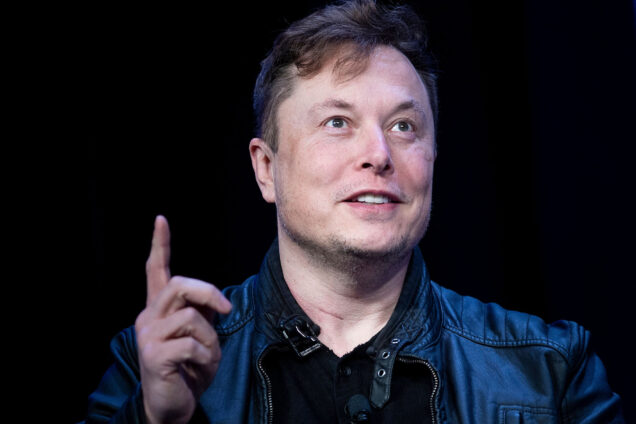 Photo: Elon Musk, a white man with thinning brown hair, looks and points up as he's shot mid-talk in front of a black background.