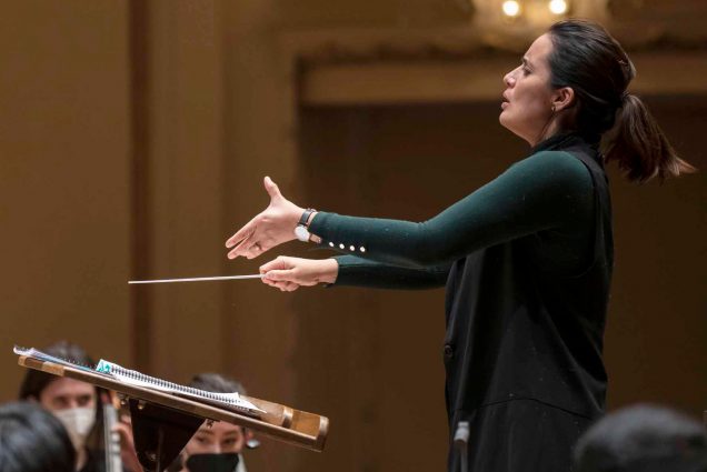 Photo: Conductor Lina Gonzalez-Granados is shown mid-performance gesturing and conducting with huge arms and movements. She wears a dark green sweater and looks expressive. Masked people sit and play various string instruments in the blurred foreground and background.