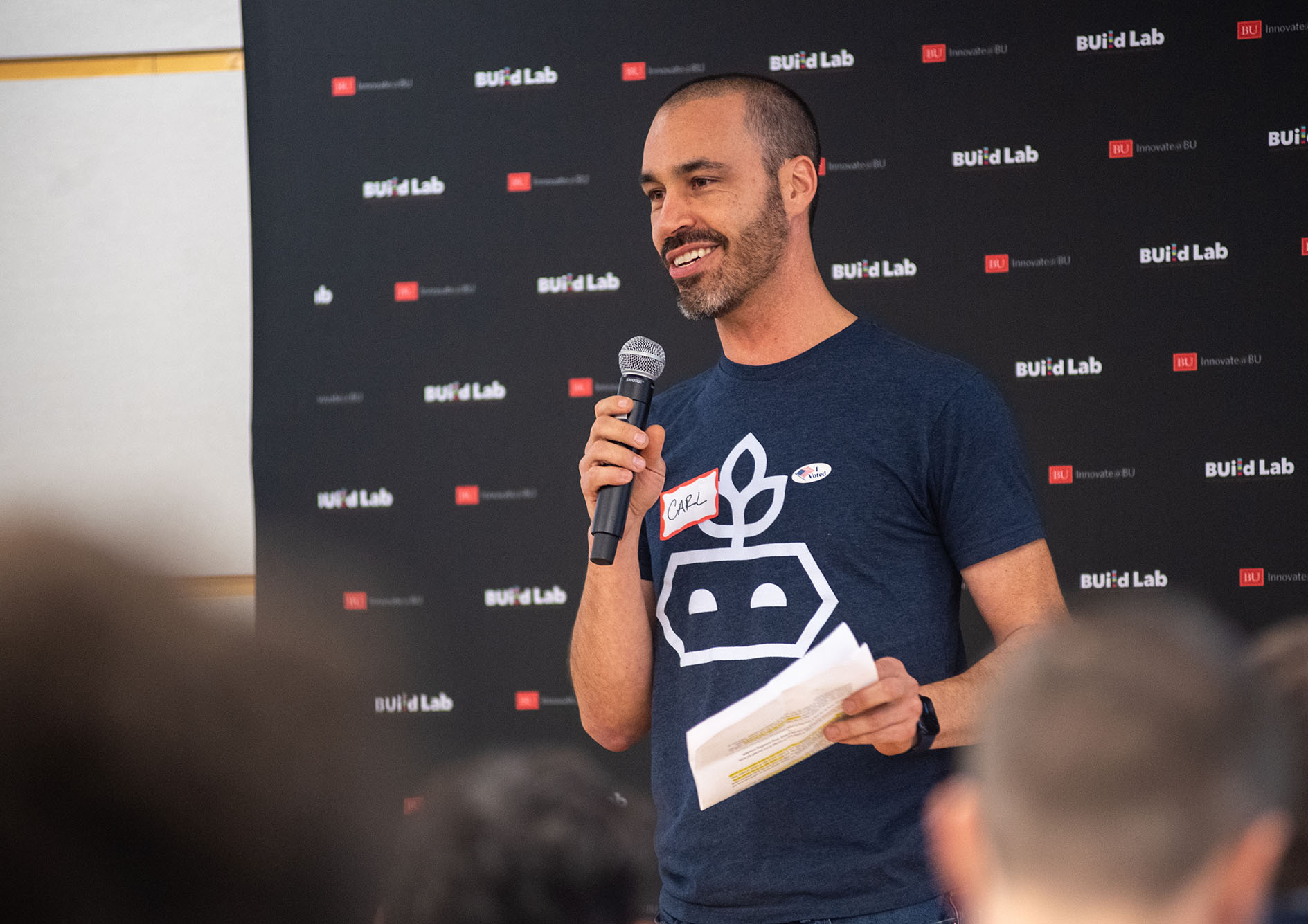 Photo: Carle Palme, a young man wearing a navy blue tee shirt, speaks into a microphone on stage with a black background featuring a repeating logo that reads "BUild Lab". 