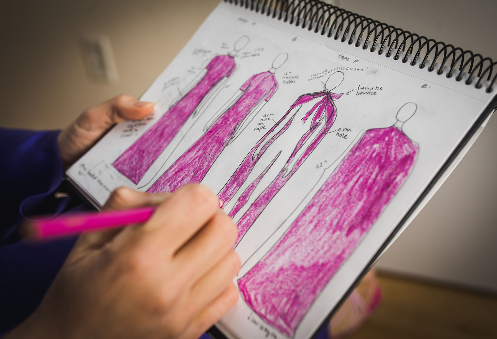 Photo: Hammouda sketching her Wendy cape and Rosemarie dress. Fuchsia pink dress is sketched on a medium-sized sketchpad as hands fill in rough drawings with the color.