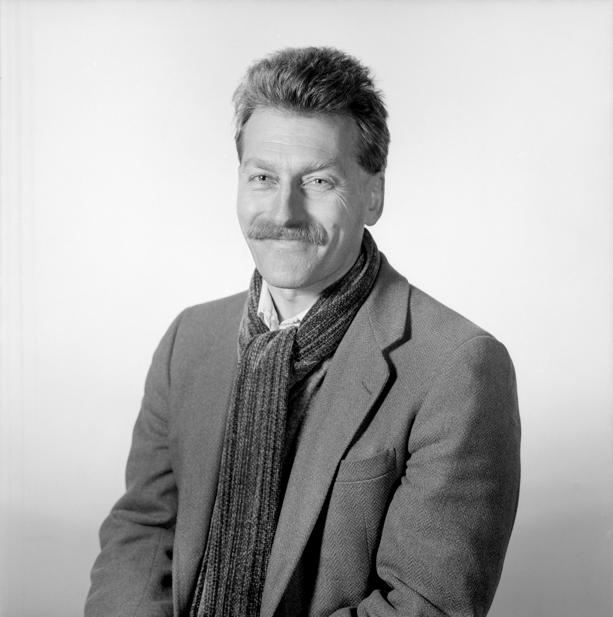 Black and white photo of Anthony Barrand. A white man with a fluffy, coifed hair sits, poses, and smiles for the camera. He wears a nightly tied scarf and blazer.