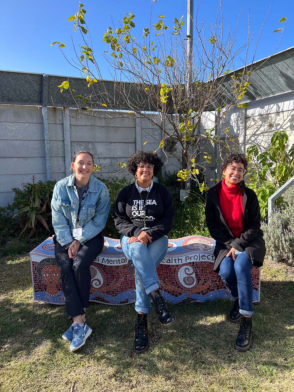 Photo: Three women smile and sit on a rock bench that reads Perinatal Mental Health Project on a sunny day.