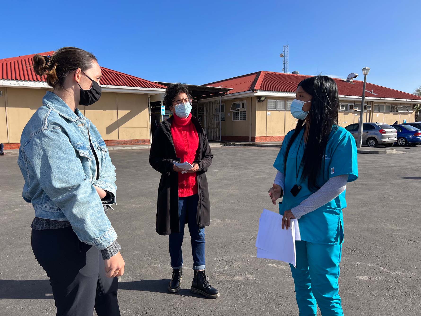 Photo: Three women talk and face each other outside a health center in South Africa. The woman on the far right wears teal scrubs and holds a few papers in her hands. All are masked.