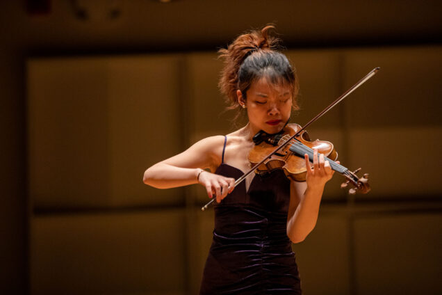 Photo: Leqing Wang (CFA’25), grand winner of the Bach Competition 2022, performs “Sonata No. 1 in G minor, BWV 1001” by Johann Sebastian Bach at Boston University’s College of Fine Arts Concert Hall on November 29. A young, tan Asian woman wearing a strappy black dress, plays a violin on stage. She looks concentrated as she holds the instrument.