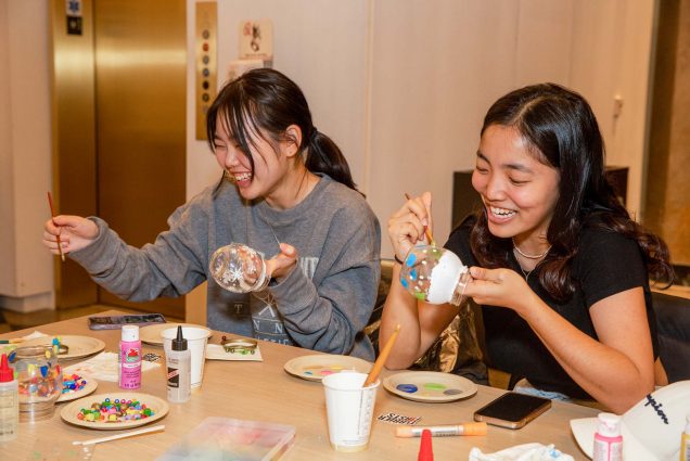 Photo: Jisu Lee (Wheelock’26), left, and Caitlyn Kyong (COM’26), right, decorate custom snow globes at the “Crafts & Chats” event within the Howard Thurman Center on the evening of November 30. Two young Asian women hold clear globes in their hands, as they laugh, paint, and decorate them.