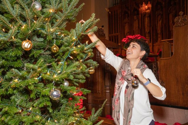 Photo: Casey Crawford, a young white person with a short hair cut and wearing a white longsleeved shirt, places one of the last few ornaments on a large fir tree before choir rehearsal starts during the annual “Hanging of the Greens” on December 8 at Marsh Chapel.