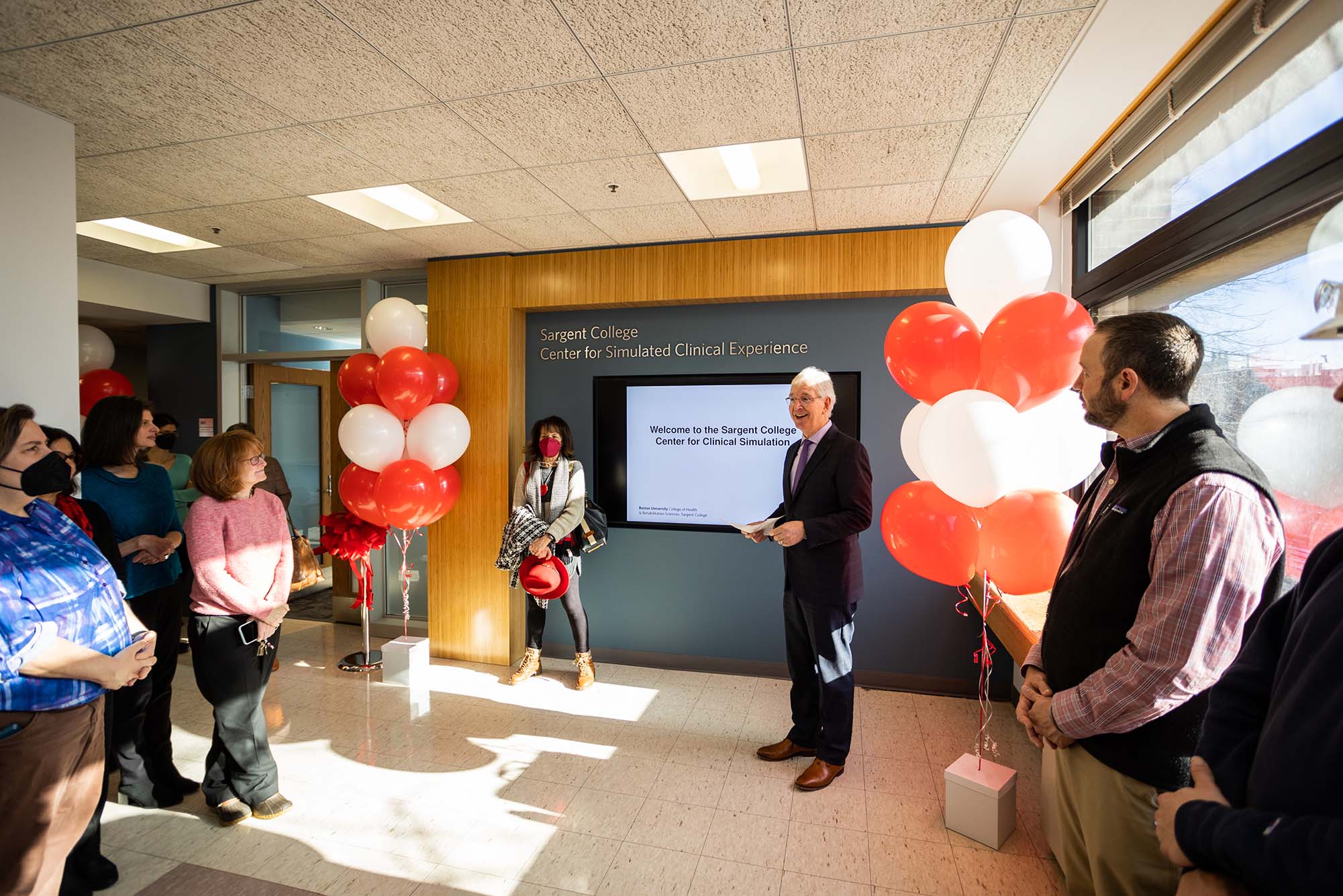 Photo: Sargent College Dean Chris Moore (center) stands in a room filled with red and white balloons to celebrate the opening of the new clinical simulation rooms. He stands in front of a dark blue wall with silver letters that read "Sargent College Center for Clinical Simulation" as well as a tv screen showing a white slide that reads the same. A small group of people stand on either side of him as they look on as he smiles and speaks.