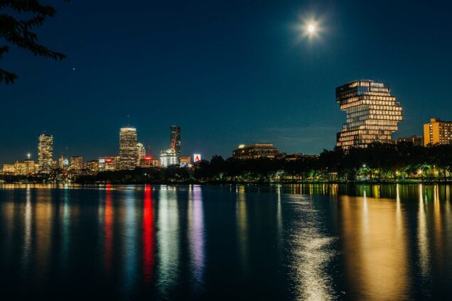 Photo: Boston University’s Center for Computing & Data Sciences is shown as part of the nighttime Boston Skyline. Buildings are lit and a reflection of the city is shown on the river in front of everything.
