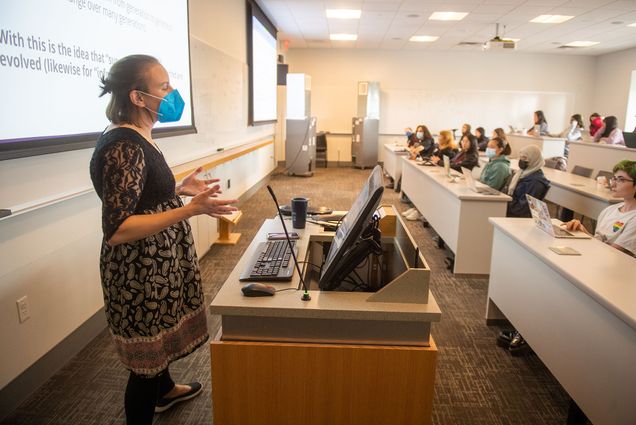 Photo: Melisa Osborne, a woman wearing a blue face mask and floral burgundy dress, leads a multi-disciplinary CAS class taking a look at the racism implicit in science and healthcare and trying to unpack it. She stands in front of a large lecture class room facing the class as she lectures.