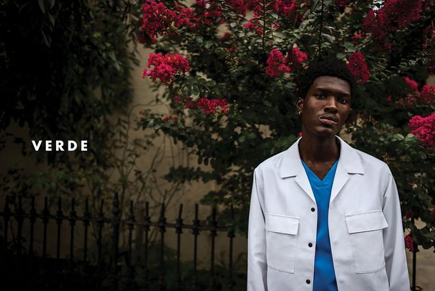Photo: A young dark-skinned Black man models blue scrubs and a white lab coat in front of a large rose bush. The name "VERDE" is shown to the left of him in bold, white letters.
