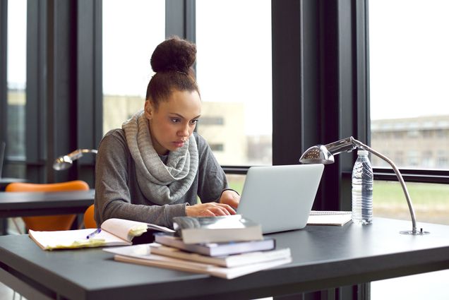 Photo: a young Black girl sits in library, typing on laptop for her assignment. She has various books and notes open in front of her on the table as she looks to her screen in concentration.