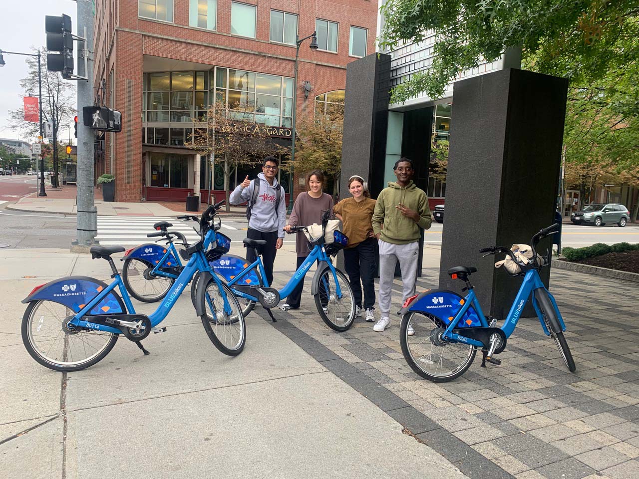 Photo: “Data harvester” team members Rishi Shah (CAS'24, MED'24) (from left), Hellarine Wu (Sargent’23), Sophie Muller (CAS’24), and Joshua Joseph (CAS’24) on a site visit in Cambridge’s Central Square in September. Four young adults smile and pose awkwardly on a large sidewalk on a gloomy day. Four blue bikes stand in front of them.