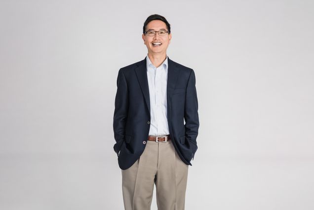 Photo of Chris Chen, an Asian American man with glasses who wears a blue suit jacket and smiles with his hands in his pockets. He stands in front of a light grey background.