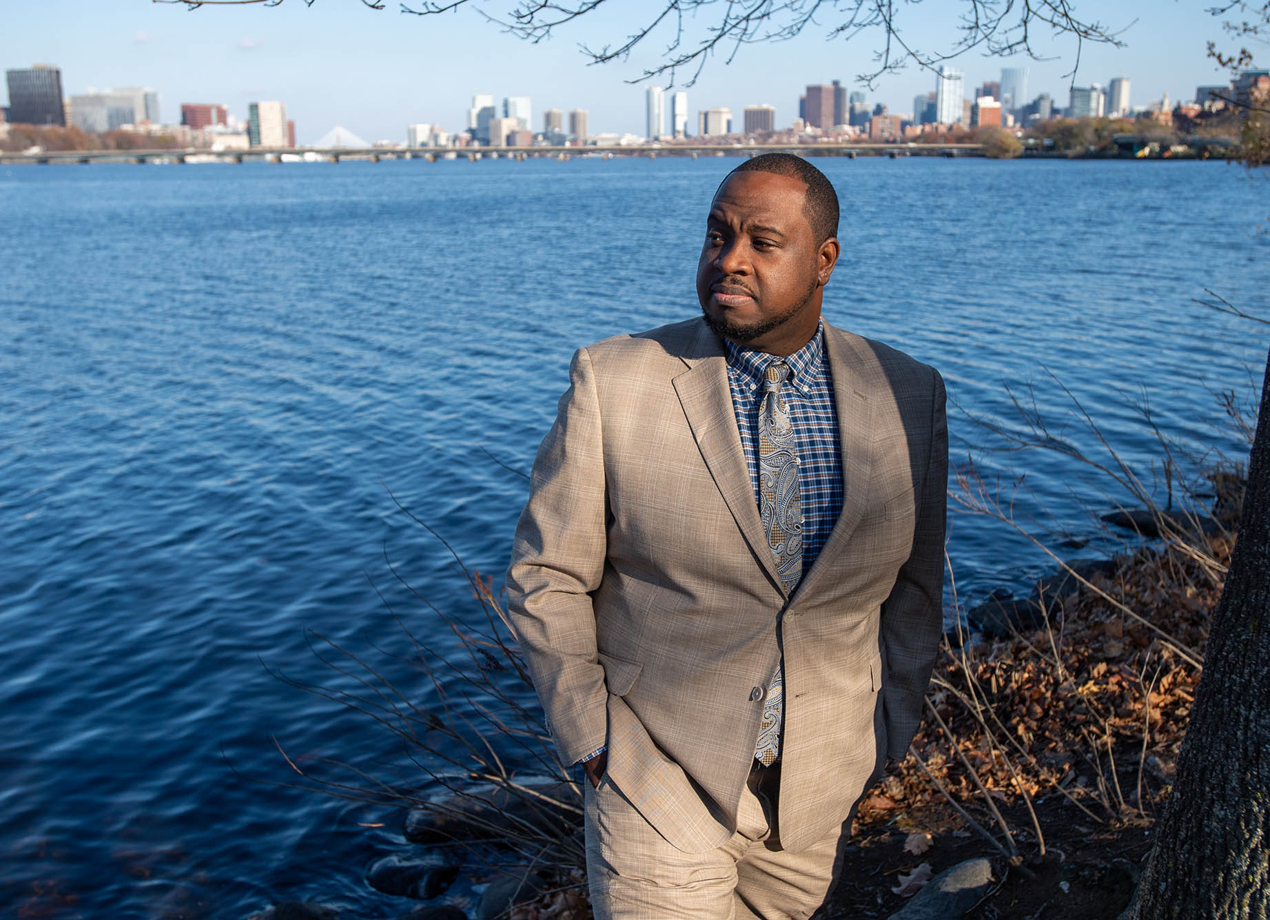 Photo: Rev. Vernon K. Walker posing along the Charles River. A Black man wearing a tan suit and light blue collared shirt looks off to the left as he poses at the bank of a large body of water. The city of Boston can be seen distantly in the background.
