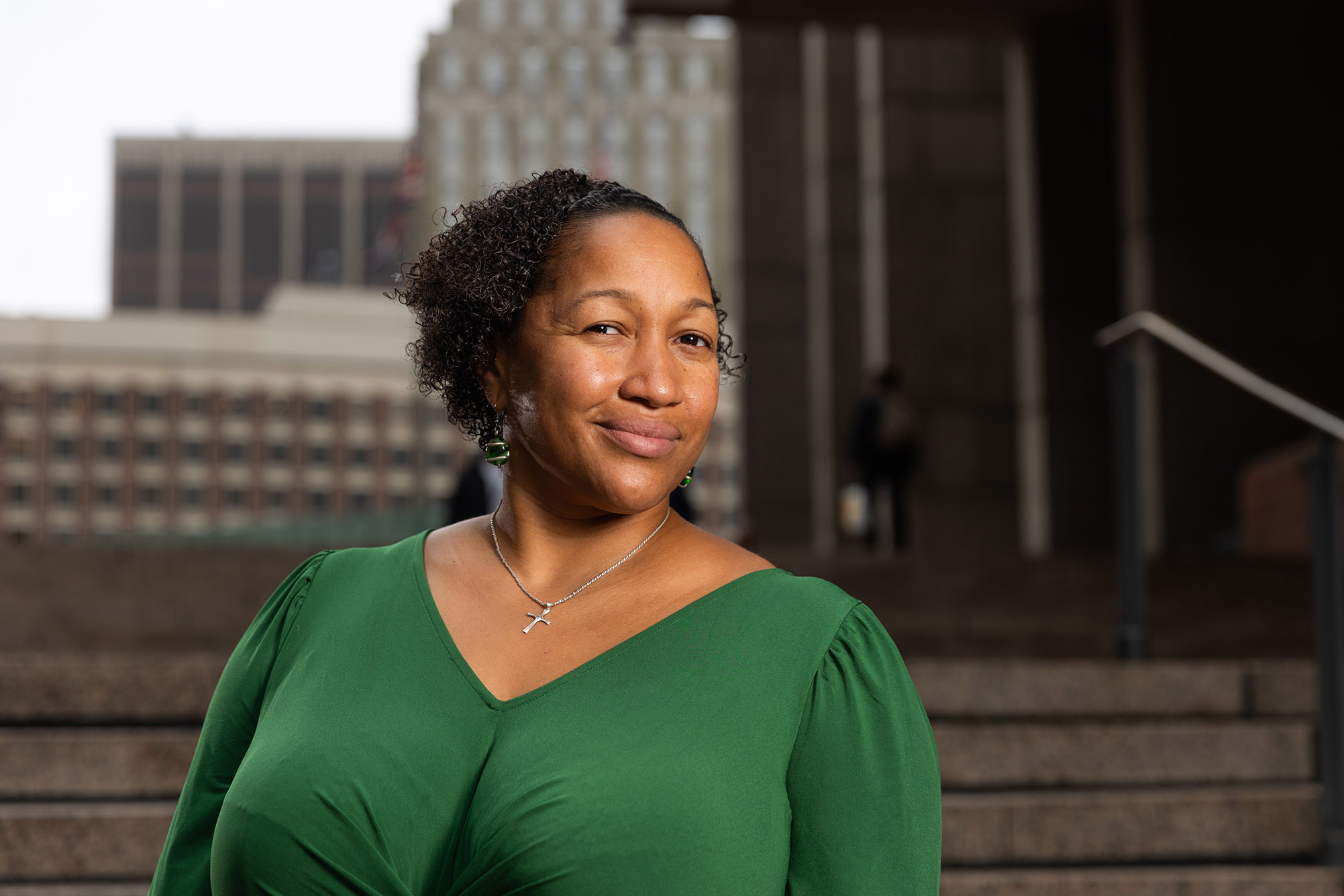 Photo: Reverend Mariama White-Hammond poses for a portrait in front of Boston City Hall. A Black woman with short, curly natural hair smiles and wears a dark green blouse. A city scape and set of cement stairs can be seen in the blurry background.