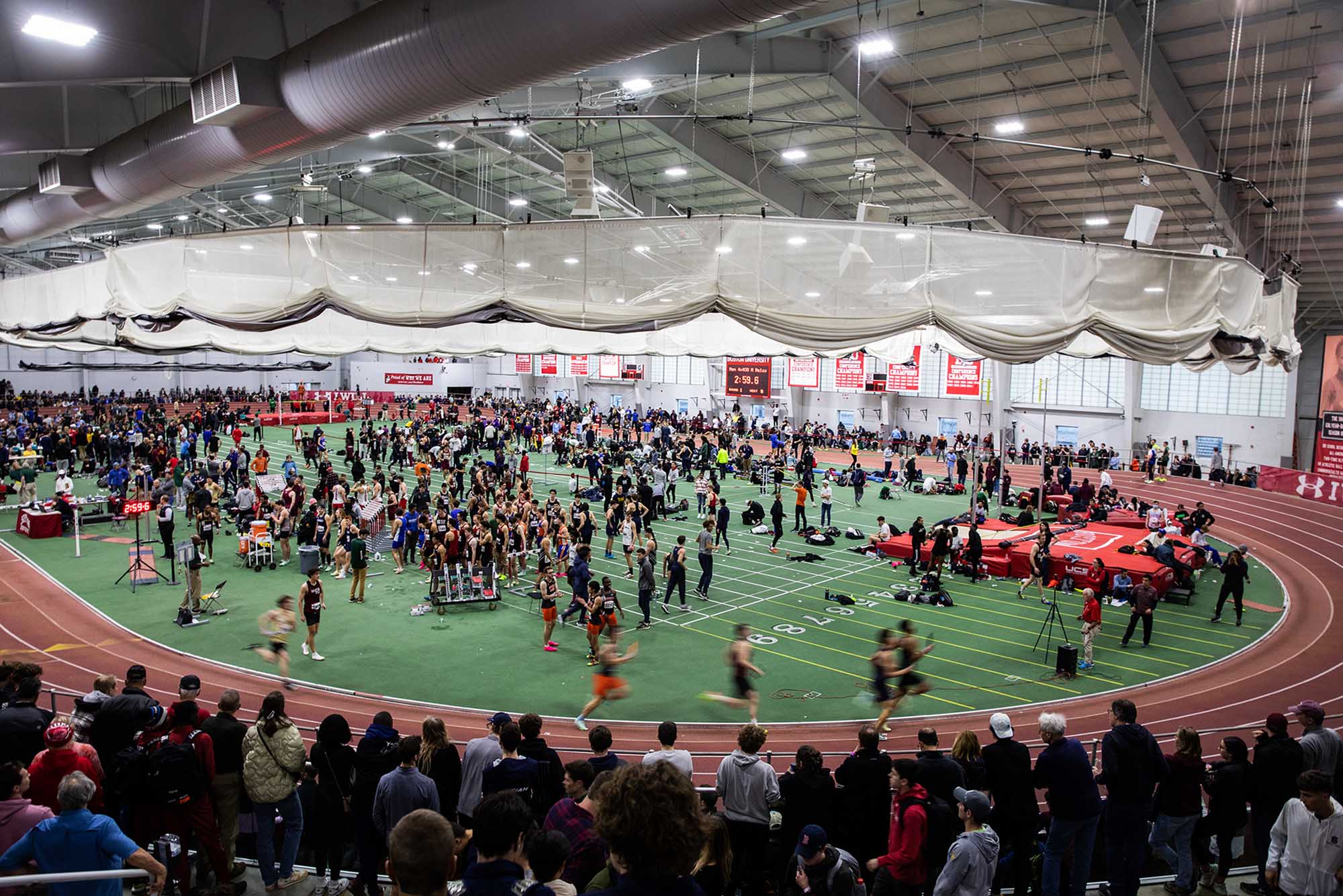 Overhead shot of the indoor track at the BU Track and Tennis Center. The shot shows bustling activity during the annual David Hemery Valentine Invitational, as runners are shown racing on the track, pole vaulters and high jumpers are on the green infield, and people spectate in the stands.