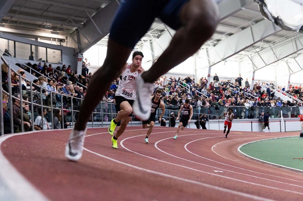 Photo: Runners sprint out on he Bu Track and tennis center's indoor oval. Five male runners from different schools, each in their own lane, sprint out at an angle on a red banked indoor track. A crowd of spectators can be seen in the stands behind them.