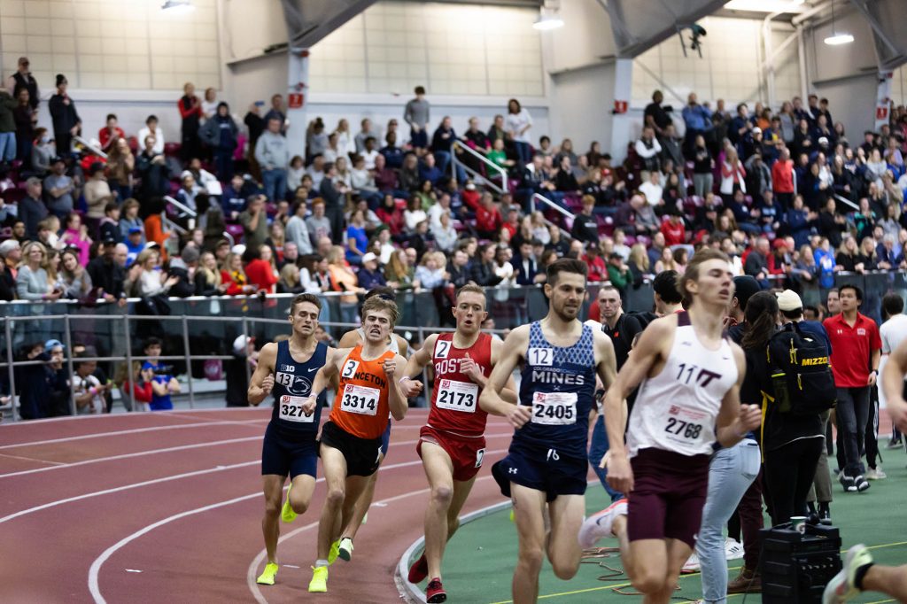 Photo: Five runners wearing jerseys from five schools: (from left) Northeastern, Princeton, BU, Colorado, and MIT, race past one of the curves on the Bu indoor track surface. A large crowd can be seen behind them in the stands.