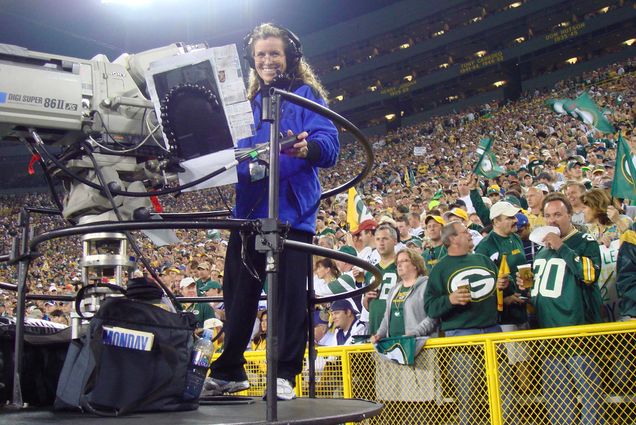 Photo: Deena Sheldon filming a Sunday Night Football game in Green Bay in 2008. A white woman with long, curly dirty blonde hair stands behind a large professional video camera on a high platform. She smiles as the photo is shot and a crowd of Green Bay Packers fans cheer in the stadium behind her.