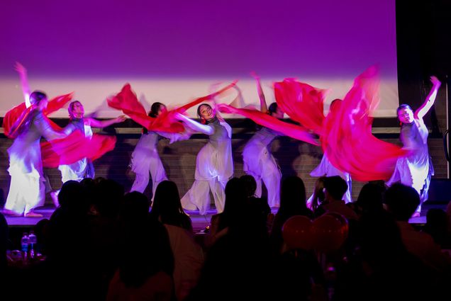 Photo: Verge, a BU traditional Chinese dance group performs at a Lunar New Year celebration hosted in the GSU ballroom Feb 3. Action shot of a group of women dressed in traditional white and grey pants and long tops dance with long red scarves on stage as an audience watches.