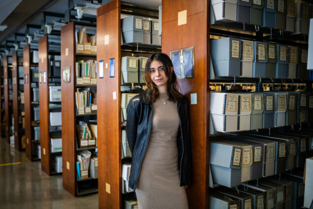Photo: Journalism student, Sahika Aydinol (COM'24) is shown posing against a stack of books in a library. A young white woman wearing a beige dress and faux leather jacket stands with hands behind back in a library/archive.