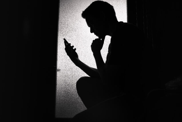 Photo: Black and white photo of a young man in shadow crouched down as he looks at a cellphone.