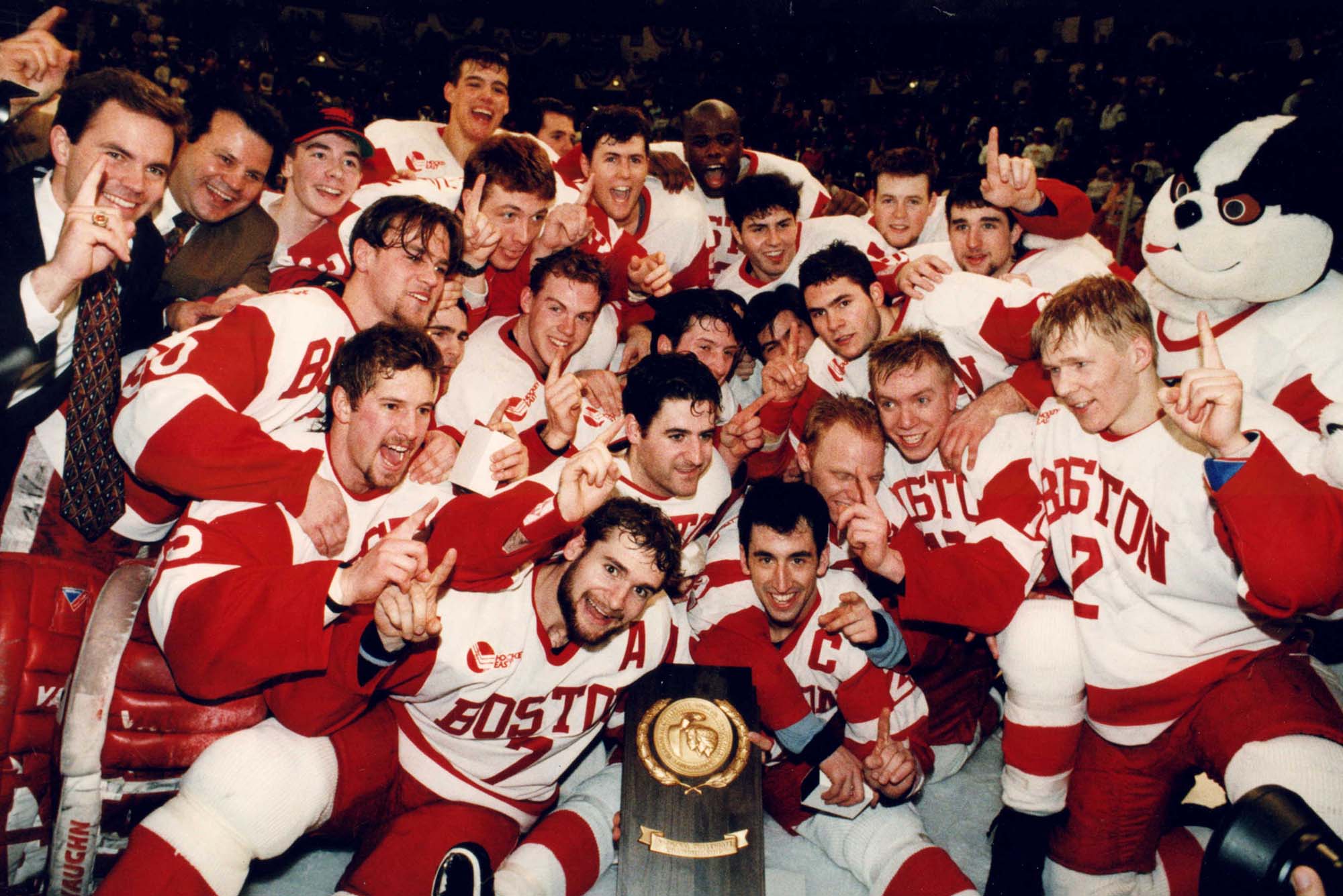 Photo: Group photo of the BU men's ice hockey team with the NCAA trophy. They all pose in the "Boston" red and white hockey uniforms and hold up a finger each signifying they're number 1.