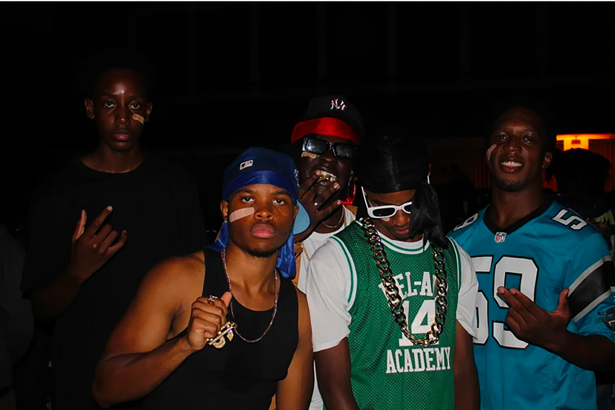 Photo: A group a Black men at an event. From left, a Black man with a black t-shirt and a bandaid on his face. In front of him, a Black man with a blue flat-cap and durag, a bandaid, a gold chain with a dollar sign pendant, and a black tank top. Behind him, a Black man with a NY Yankees flat-cap, a red durag, black sunglasses, and a white t-shirt. In front of him, a Black man with a black durag, a green jersey over a white t-shirt, a thick gold chain, and white sun glasses. He is looking down. And on the far right, a Black man with a light blue jersey poses, smiling.