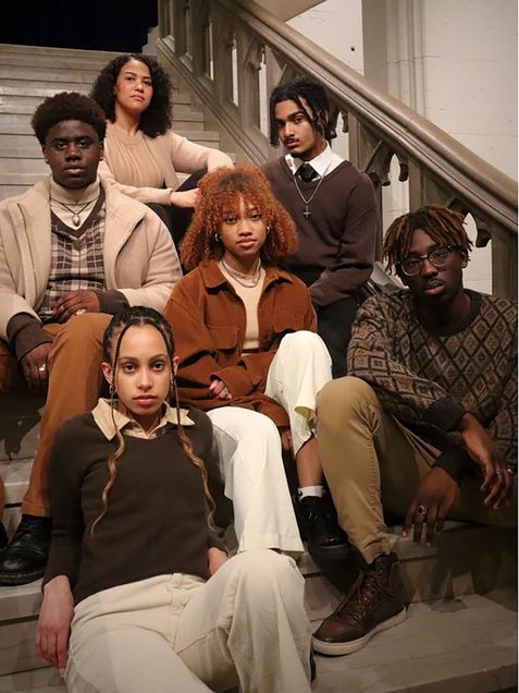 Photo: A group of Black individuals on a staircase. From bottom left, a feminine-presenting individual with tighter box braids sits in front. They are wearing a monochrome brown outfit. Behind them, to the left, sits a masculine-presenting individual with a similar color palette on. They have short hair, curly and combed out that sits tall on top of their head. Behind them, to the center, sits a feminine-presenting individual with short, shoulder length curly hair in the same monochrome color scheme. In front of them in the center of the group and photo, sits a feminine-presenting individual with shoulder-length, auburn-dyed hair. They are wearing a burnt-orange shacket and white pants. Behind them, on the right, a masculine-presenting individual with chin length locs wears a darker brown sweater vest. In front of them, in the bottom right-hand corner of the photo sits a masculine-presenting individual with a grandpa-esque sweater in the muted brown color palette the rest of the group wears. They have circular glasses and eye-length, partly dyed locs.