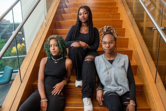 Photo: Three Black women sit on an orange-brown staircase. From left, the women has blue-green shoulder-length locs. She is wearing an all black outfit. In the middle, a women with long, black box braids wears an all black outfit. And finally, on the right, a women with a blonde ombre locs that are tied up in a high pony. She wears an all black outfit with a heather gray, oversized sweater vest overtop and glasses that match the orange-brown of the stairs.