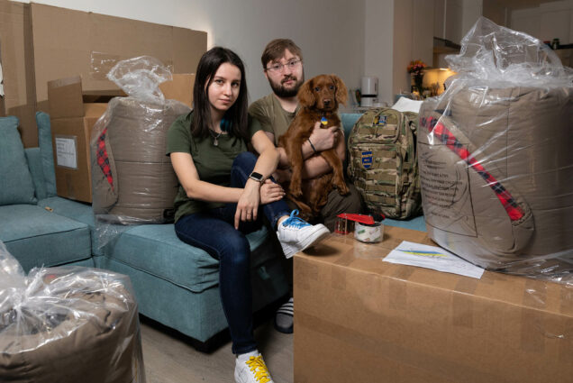 Photo: A couple sits with their dog amongst various packed boxes and war supplies for Ukrainian individuals. They are wearing neutral clothes and are looking straight into the camera.