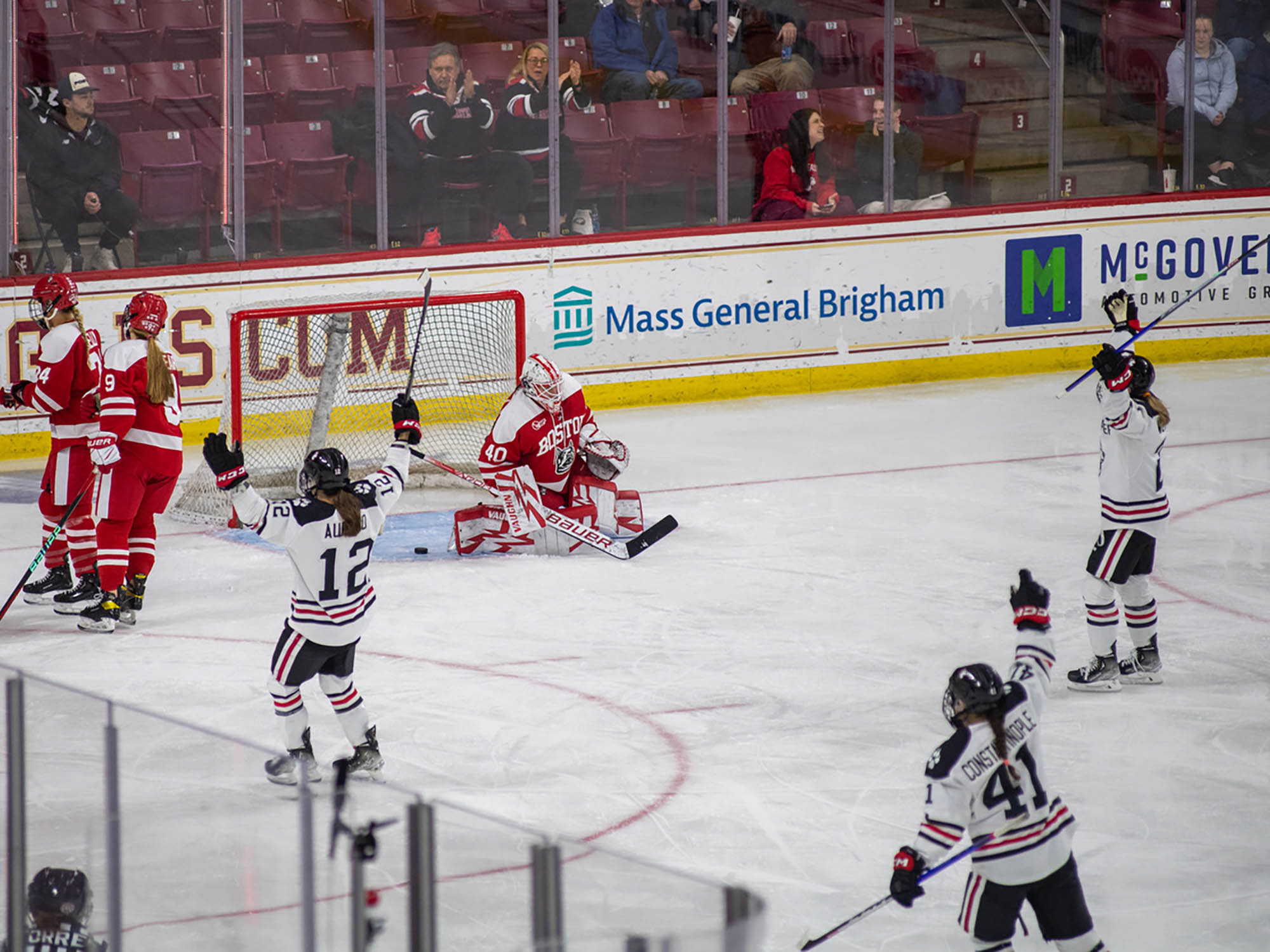 Photo: NE Huskies celebrate their goal on an ice rink in their navy and white uniforms. The BU Terrier team is in all red with white detailing uniforms. They are at the foreground of the photo. In the background, behind the protective glass, spectators of the game sit sporadically in the left area of the photo.