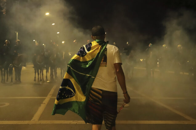 Photo: Bolsonarista terrorists generate chaos in Brasilia, Brazil in a coup attempt with the invasion of the STF, National Congress and Palacio do Planalto. Seen from the back, A Braizilian person wearing the Brazilian flag as a cape stands and faces a line of armed officers in riot gear. Scene is at night, as a fog is seen in the air.