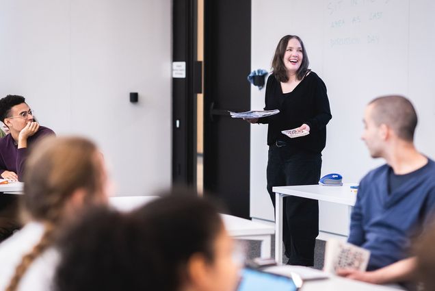 Photo: Professor Amy Appleford leads an ENG 101 Encounters class at the Center for Computing & Data Science. A white woman with brown hair and wearing a black long sleeved top and pants, laughs and looks to the right as she holds a stack of handouts. She stands in front of a bright classroom as student sit in seats in front of her and turn to look at her.