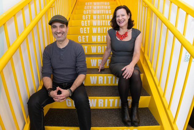Photo: Artistic director Dariel Suarez (GRS’12) (left) and founder and executive director Eve Bridburg (GRS’97) sit and pose on a set of bright yellow stairs. Each step has the words "Get writing" printed on them in white. A latino man wearing a grey beret, grey shirt, and black pants sits and smiles with hands clasped in front of him. To his right, a white woman wearing a grey dress, black pantyhose, and red chunky necklace smiles and sits with one hand in lap and one on the stair.