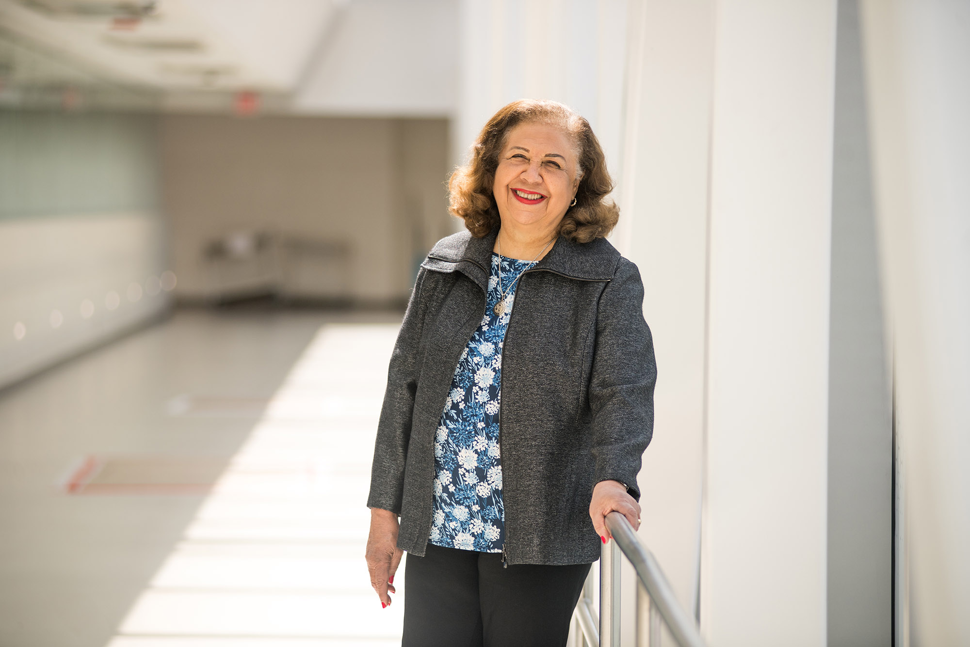 Photo: Marcelle Willock, MD. A light-skinned, older Black woman with light brown hair smiles and poses in the hallway of a bright, sunlit hospital. She wears a blue floral blouse, grey jacket, and black pants.