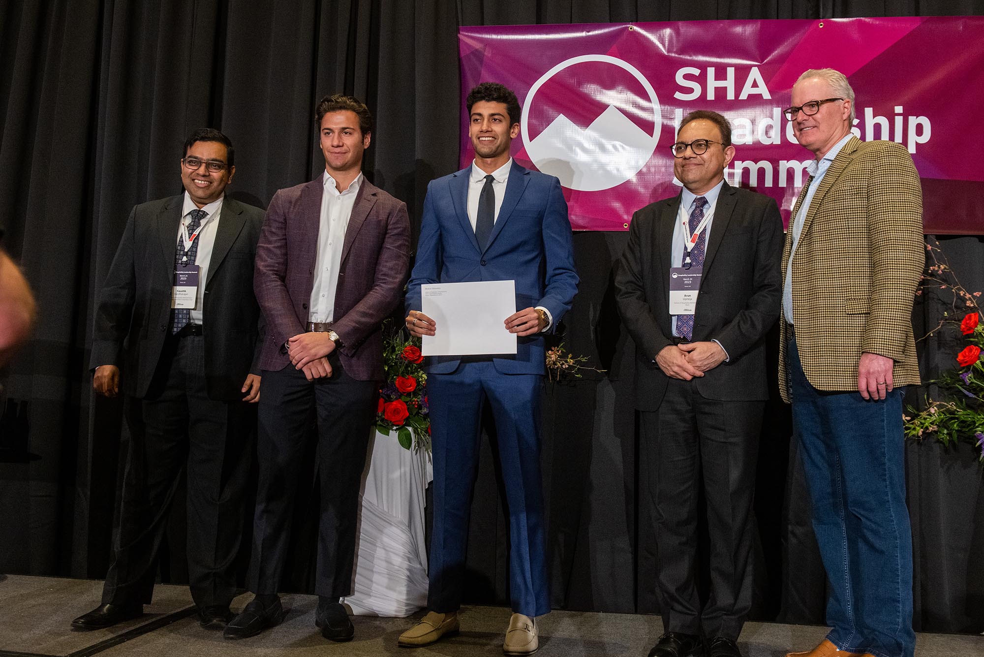 Photo: Five men stand on a stage to celebrate the winner of a compeition. From left, a West Asian man wearing a black suit, a white man in a maroon blazer and black pants, a West Asian man wearing a blue suit with a black tie, an older West Asian man wearing a black suit with a purple tie, and a white man wearing a plaid blazer with tailored jeans and glasses. They are all smiling for the photo, looking to the left at another camera.