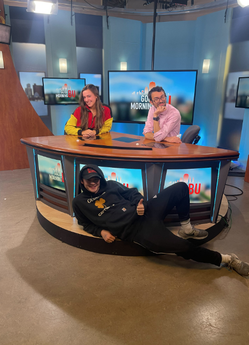 Senior, Kelsey Martirano (COM'23), pictured in the BUTV10 news room, accompanied by two other students. One student is smiling to the right of her in a chair and the other lying below the news desk, giving a thumbs up.
Picture credit: Kelsey Martirano (COM'23)