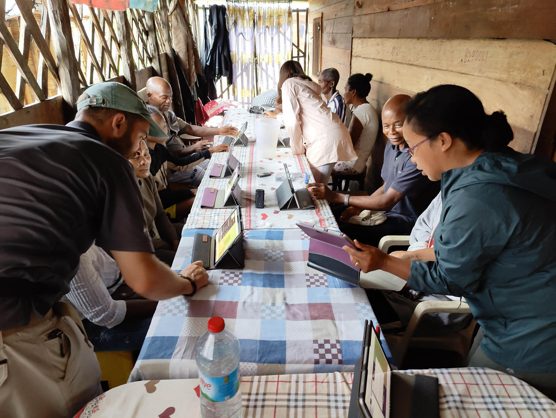 Photo: To the far left, Andrew Reid Bell, a white man wearing a green cap, looks on as a small group of dark-skinned Black women and men look to tablets on a multi-colored, checkered long table. They are in a small wooden dwelling with an open doorway and cross-patterned open windows letting light in. Amidst them, other researchers help people with their tablets.