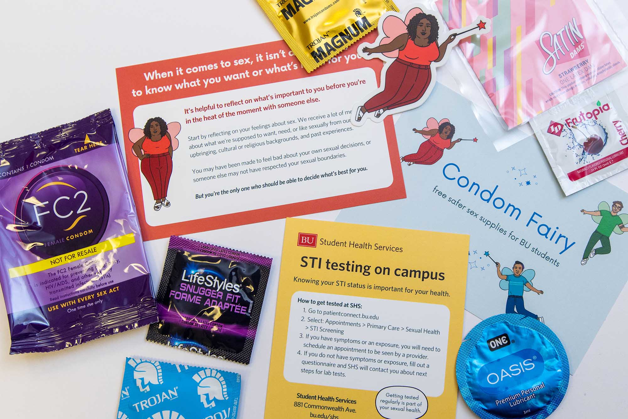 Photo: A flat-lay of the supplies found when one orders from the Condom Fairy program located at BU. Various information cards and condoms are scattered on a white table. A featured image of the condom fairy characters in sticker form lay on top of the various cards.