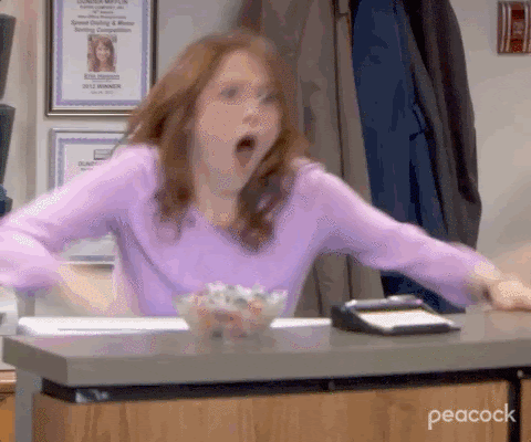 This is a GIF of a woman in an office building. The woman is wearing a purple shirt an has red hair and the GIF shows her standing up quickly from her desk cheering. She is fist-pumping her hand in the air out of excitement.