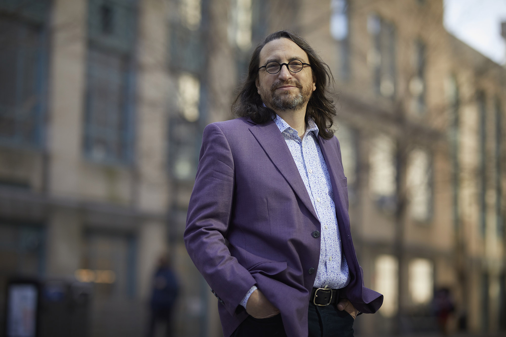 Photo: Jay Wexler, a white man with shoulder length dark brown hair and a short beard, poses with hands in pockets. He wears glasses, a purple blazer, and light blue collared shirt and stands in front of a blurry city background.