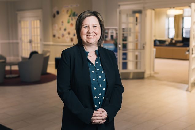 Photo: Melissa Holt, a white woman with shoulder length straight brown hair ad wearing a blue blouse with white polka dots and a black blazer smiles and poses with hands clasped in front of her. Holt poses for a portrait inside the lobby of Kilachand Hall.