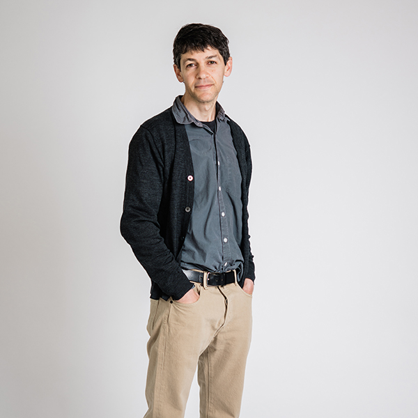 Photo: Ray Fisman, a white man wearing a dark teal collared shirt, black cardigan, and tan slacks, poses with hands in pockets in front of a white background.