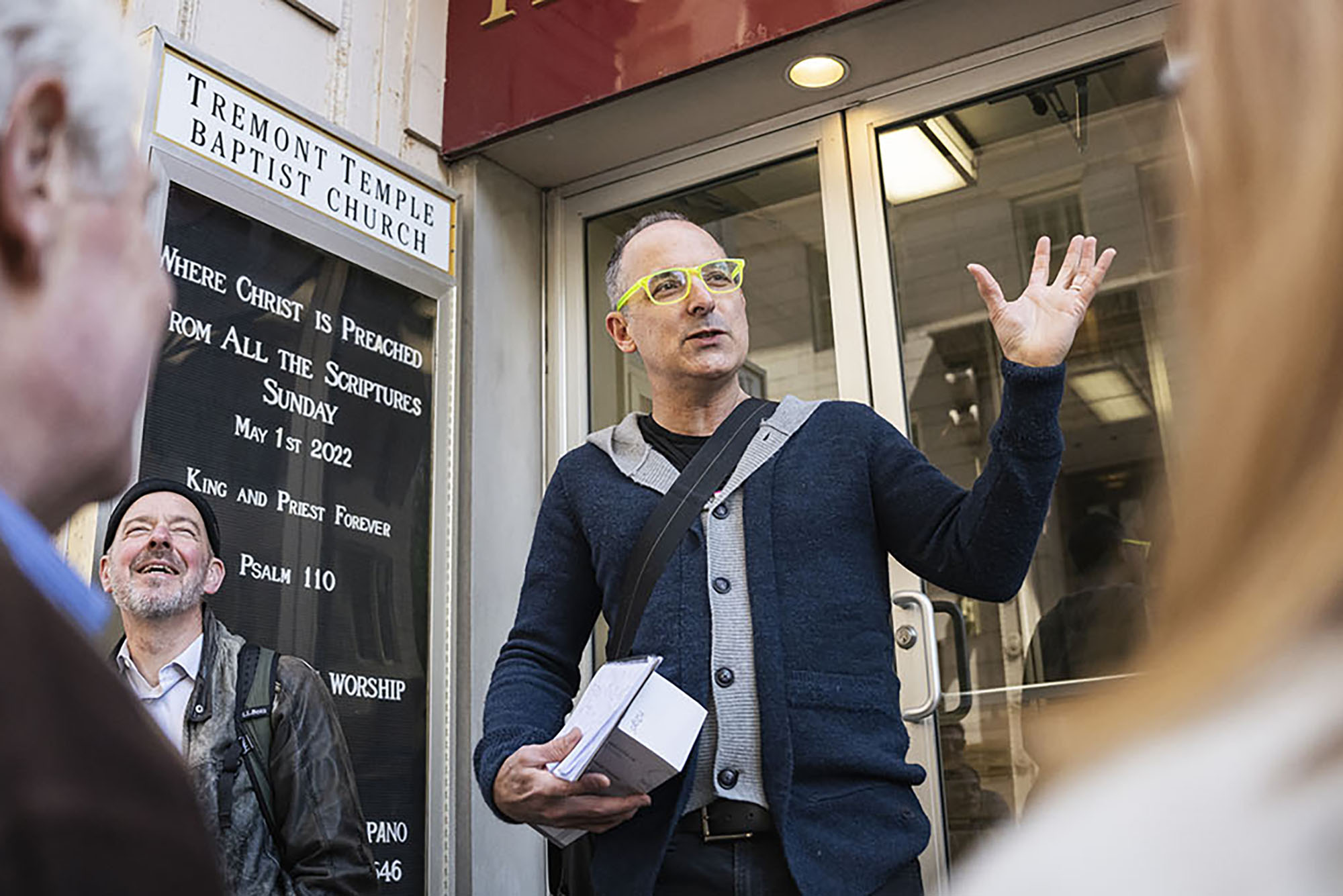 Photo: Journalist Scott Kirsner, a white man wearing neon yellow glasses, a black shirt, navy blue and grey button-up sweater, and jeans holds his hand up as he talks to a crowd. He clutches a few miscellaneous items in his other hand as he stands in front of glass doors to a building. The sign to his left reads "Tremont Temple Baptist church". Another man to his left smiles and laughs.